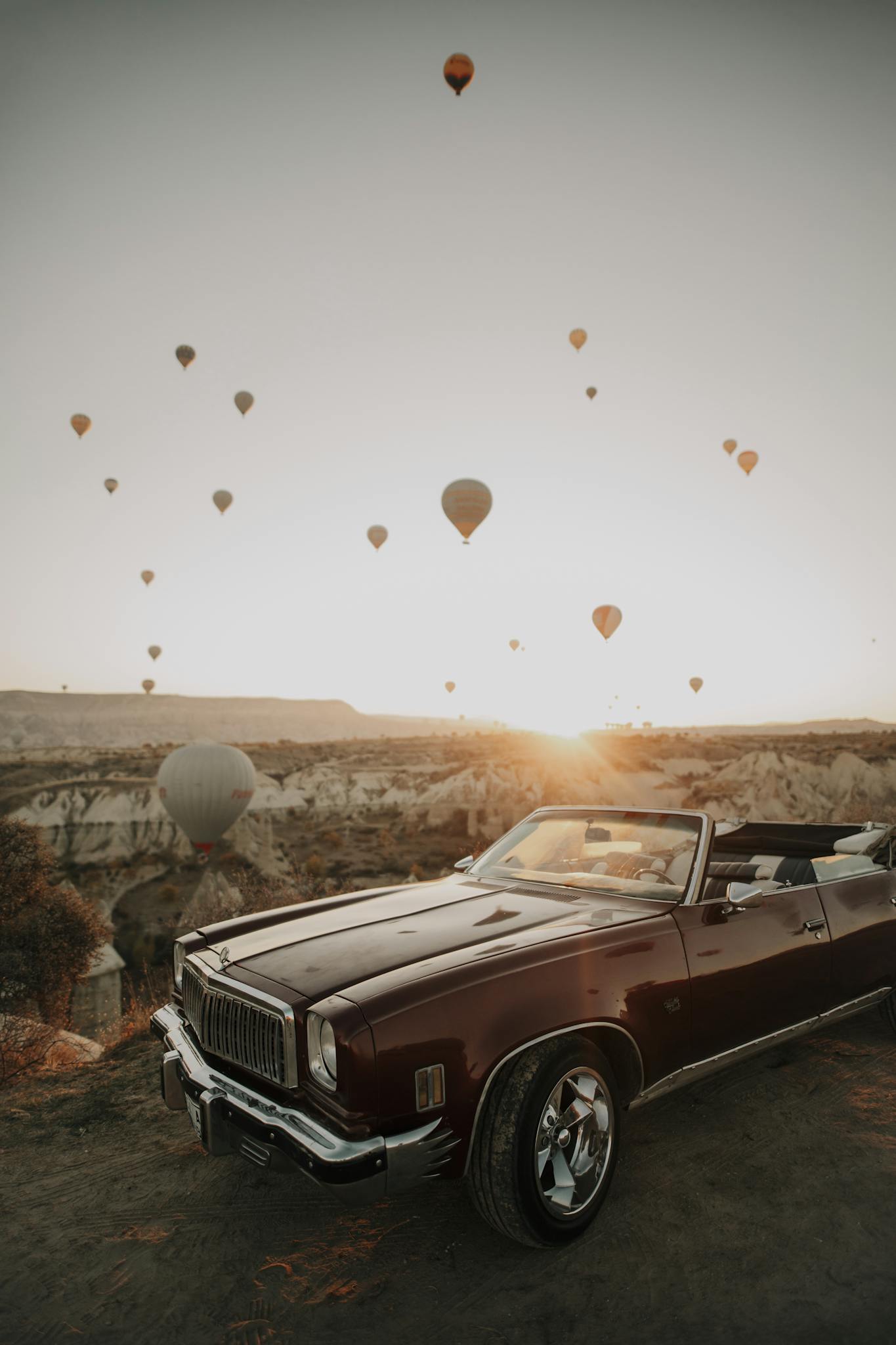 A vintage convertible car with hot air balloons at sunrise in Cappadocia, Türkiye.