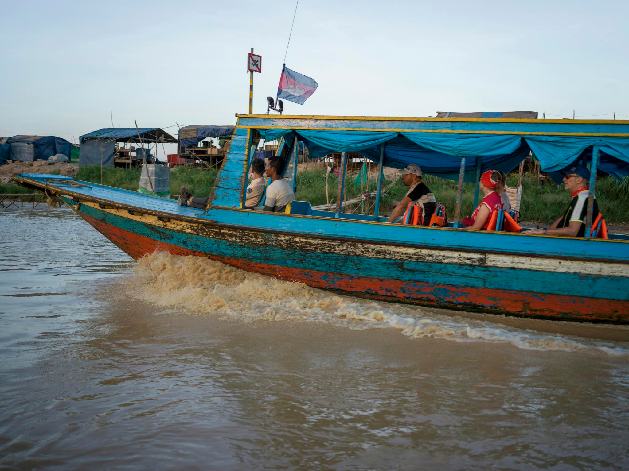 Colorful motorboat carrying people through a rural Cambodian river scene.